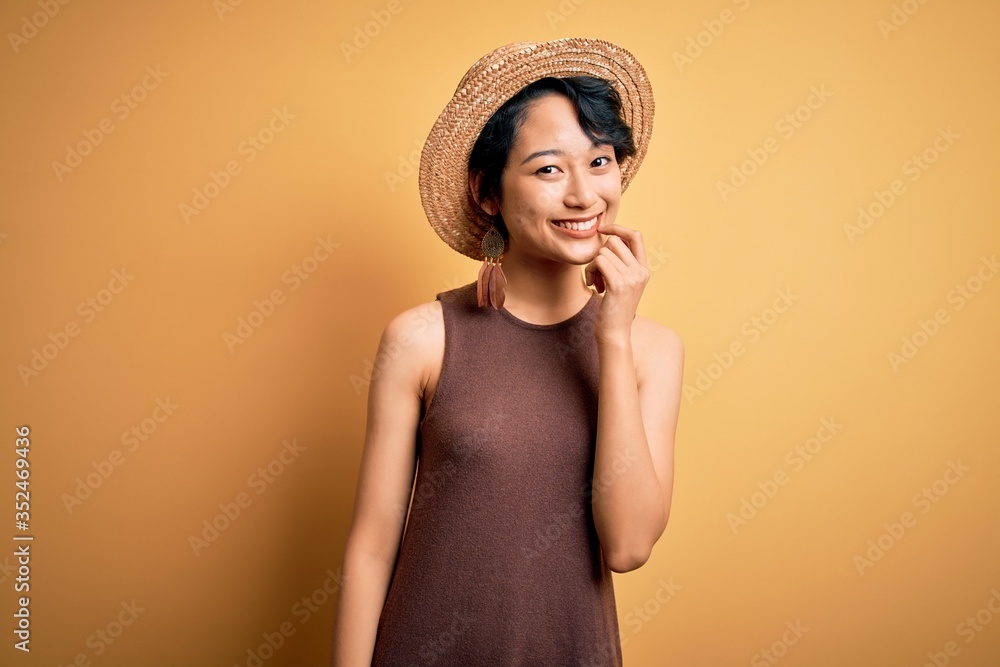 Young beautiful asian girl wearing casual t-shirt and hat over isolated yellow background looking confident at the camera with smile with crossed arms and hand raised on chin. Thinking positive. Young beautiful asian girl wearing casual t-shirt and hat over isolated yellow background looking confident at the camera with smile with crossed arms and hand raised on chin. Thinking positive.