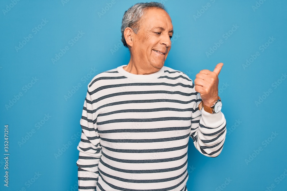 Naklejka premium Senior handsome grey-haired man wearing striped sweater over isolated blue background smiling with happy face looking and pointing to the side with thumb up.