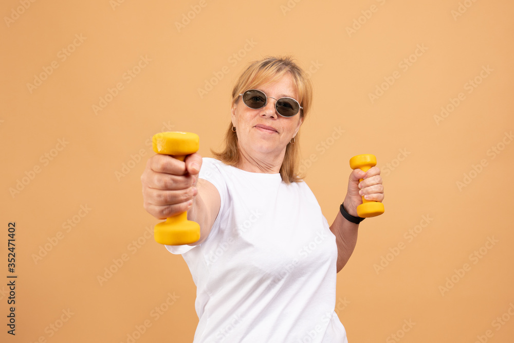 Excited senior fitness woman in sunglasses training with dumbbells isolated on orange Excited senior fitness woman in sunglasses training with dumbbells isolated on orange