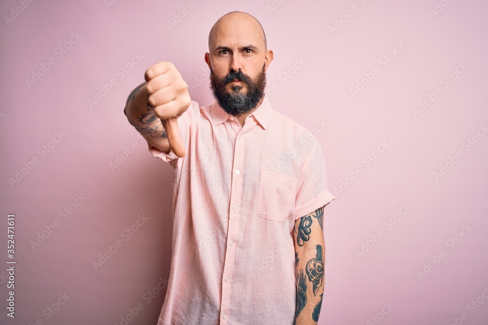 Handsome bald man with beard and tattoo wearing casual shirt over isolated pink background looking unhappy and angry showing rejection and negative with thumbs down gesture. Bad expression. Handsome bald man with beard and tattoo wearing casual shirt over isolated pink background looking unhappy and angry showing rejection and negative with thumbs down gesture. Bad expression.