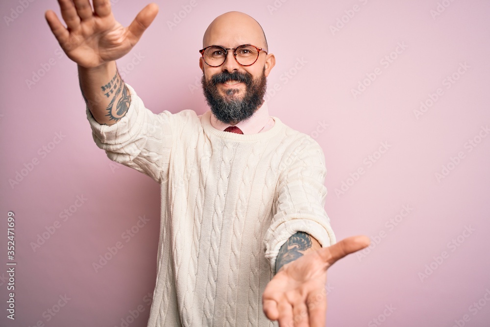 Handsome bald man with beard and tattoo wearing glasses and sweater over pink background looking at the camera smiling with open arms for hug. Cheerful expression embracing happiness. Handsome bald man with beard and tattoo wearing glasses and sweater over pink background looking at the camera smiling with open arms for hug. Cheerful expression embracing happiness.