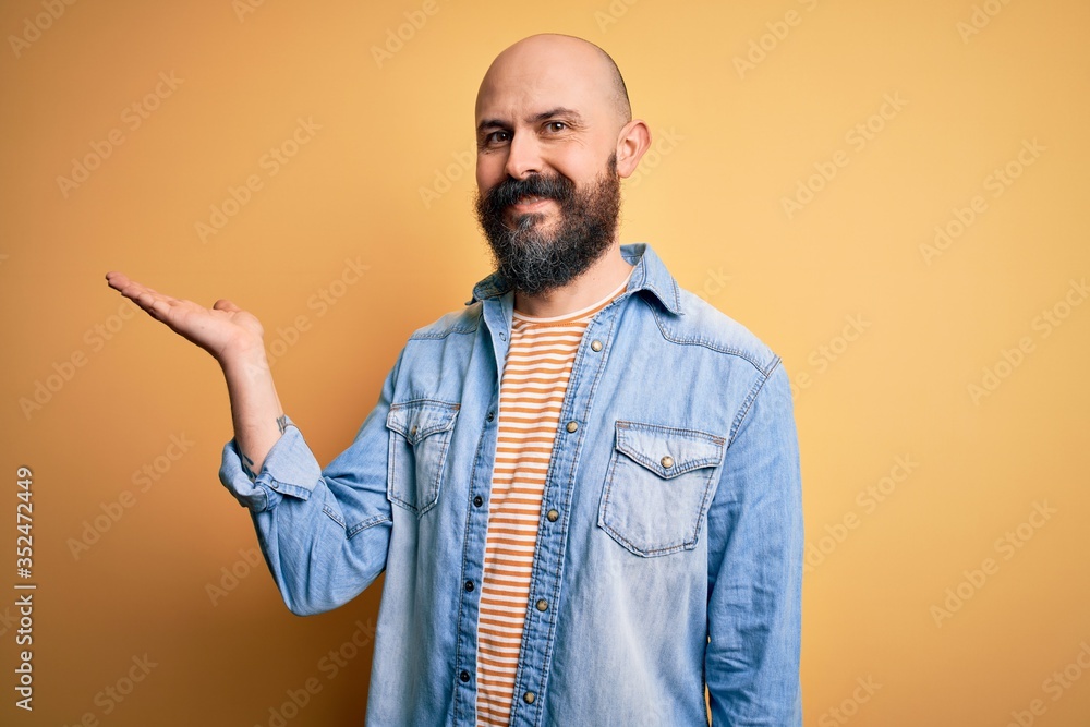 Handsome bald man with beard wearing casual denim jacket and striped t-shirt smiling cheerful presenting and pointing with palm of hand looking at the camera. Handsome bald man with beard wearing casual denim jacket and striped t-shirt smiling cheerful presenting and pointing with palm of hand looking at the camera.