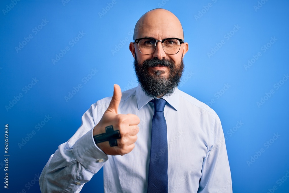 Handsome business bald man with beard wearing elegant tie and glasses over blue background doing happy thumbs up gesture with hand. Approving expression looking at the camera showing success. Handsome business bald man with beard wearing elegant tie and glasses over blue background doing happy thumbs up gesture with hand. Approving expression looking at the camera showing success.
