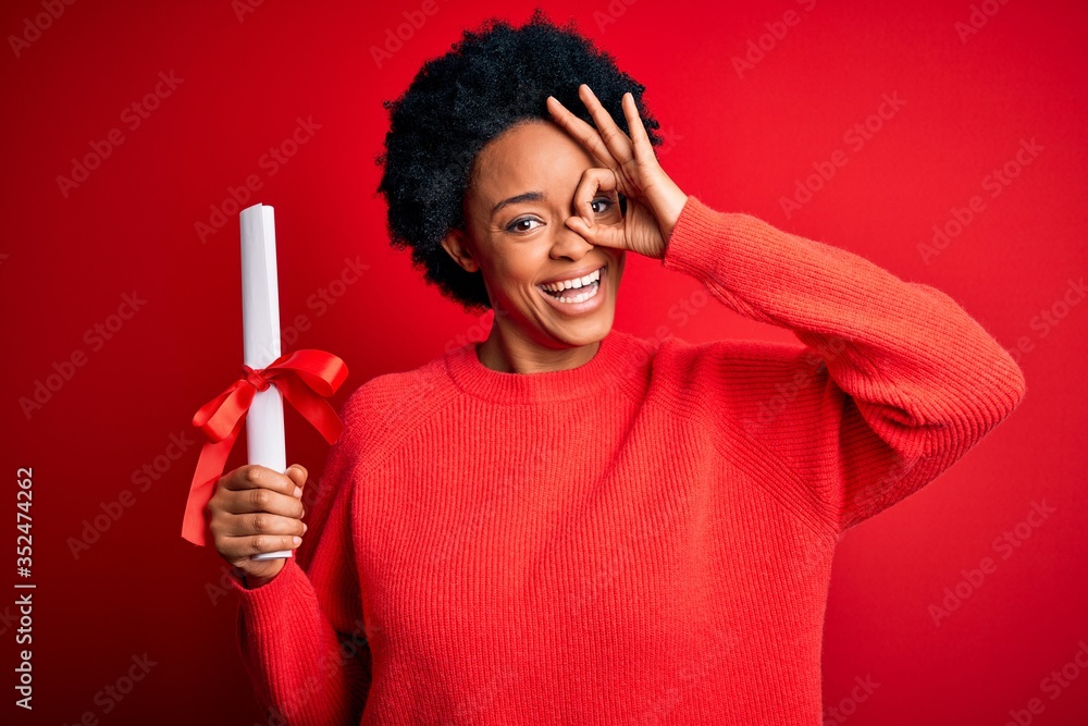 Young African American afro student woman with curly hair holding university degree diploma with happy face smiling doing ok sign with hand on eye looking through fingers Young African American afro student woman with curly hair holding university degree diploma with happy face smiling doing ok sign with hand on eye looking through fingers