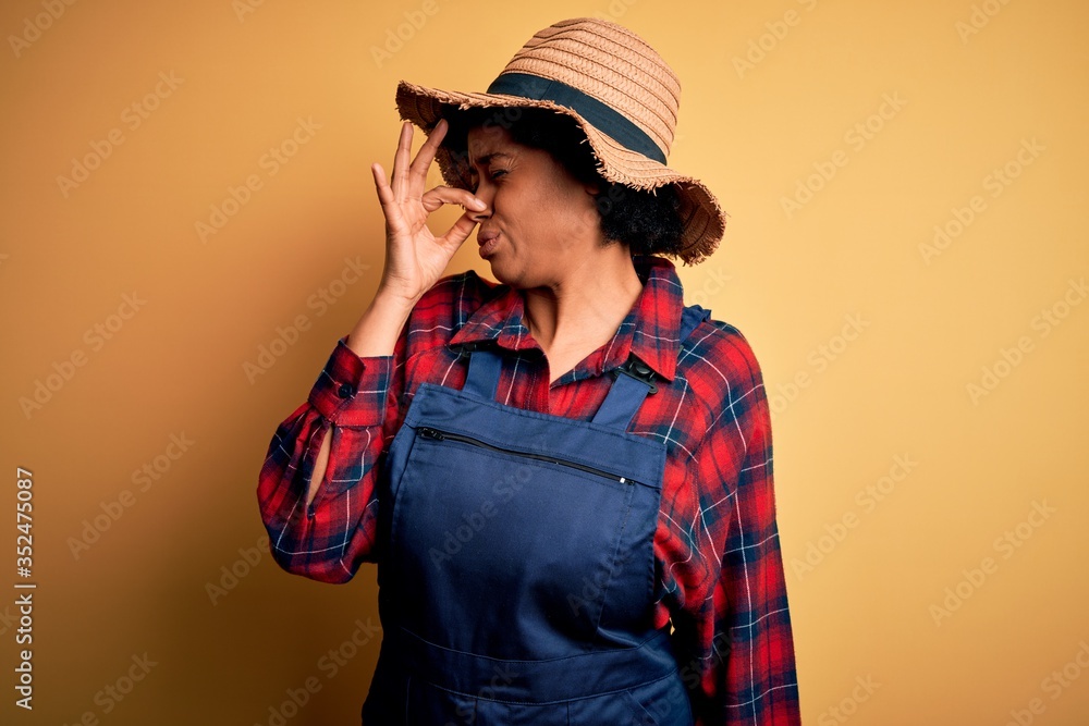 Young African American afro farmer woman with curly hair wearing apron and hat smelling something stinky and disgusting, intolerable smell, holding breath with fingers on nose. Bad smell Young African American afro farmer woman with curly hair wearing apron and hat smelling something stinky and disgusting, intolerable smell, holding breath with fingers on nose. Bad smell