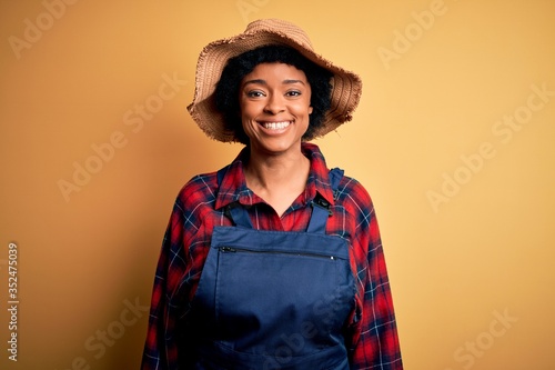 Young African American afro farmer woman with curly hair wearing apron and hat with a happy and cool smile on face. Lucky person.