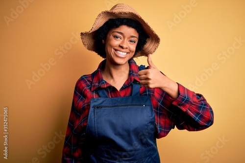 Young African American afro farmer woman with curly hair wearing apron and hat doing happy thumbs up gesture with hand. Approving expression looking at the camera showing success.
