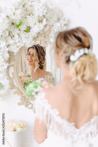 Preparing for the wedding. Beautiful young bride in white wedding dress in the room. A luxury model looks at her reflection in the mirror, with a bouquet of flowers, as if at home in a Studio room .