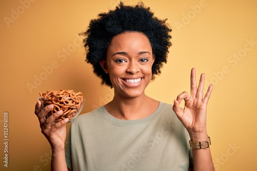Young African American afro woman with curly hair holding bowl with baked German pretzels doing ok sign with fingers  excellent symbol