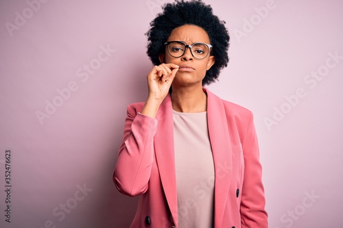 Young beautiful African American afro businesswoman with curly hair wearing pink jacket mouth and lips shut as zip with fingers. Secret and silent, taboo talking