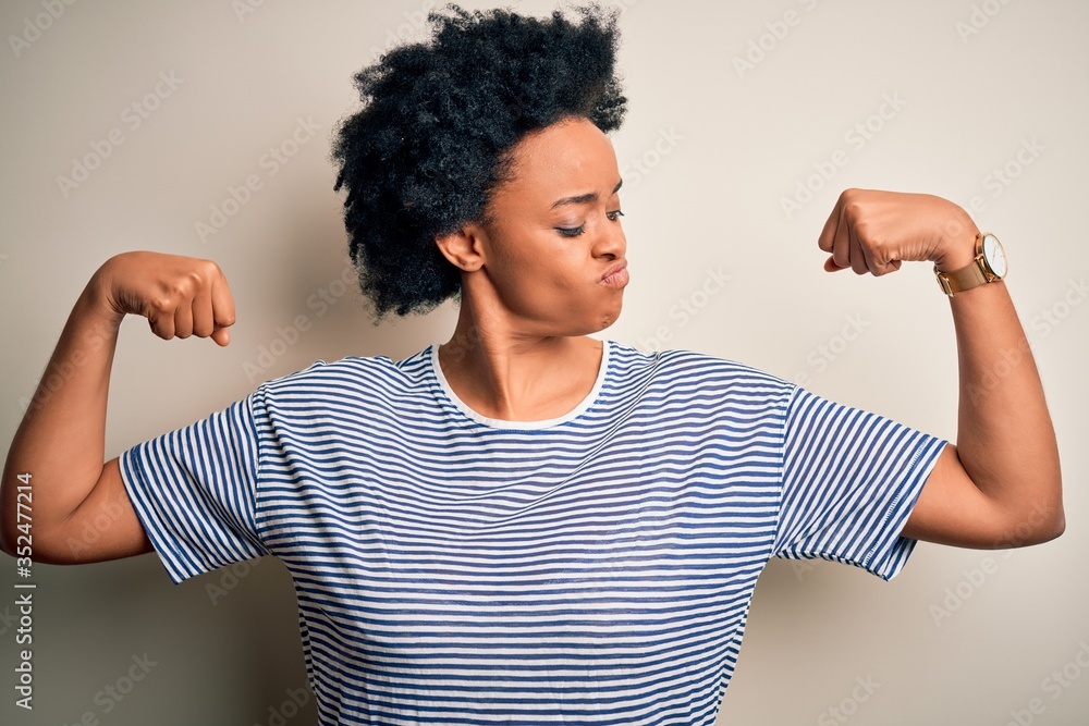 Young beautiful African American afro woman with curly hair wearing striped t-shirt showing arms muscles smiling proud. Fitness concept. Young beautiful African American afro woman with curly hair wearing striped t-shirt showing arms muscles smiling proud. Fitness concept.