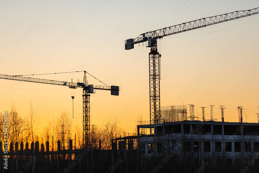 Industrial construction cranes and buildings silhouettes at a construction site on the background at sunset or dawn.