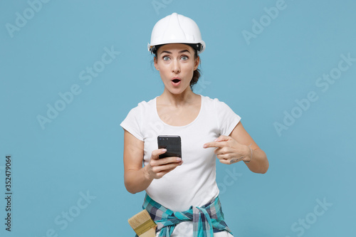 Amazed young woman in protective helmet hardhat pointing index finger on mobile phone isolated on blue background studio. Instruments accessories for renovation apartment room. Repair home concept.