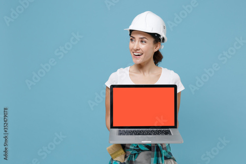Funny woman in helmet hardhat hold laptop pc computer with blank empty screen isolated on blue background. Instruments accessories for renovation apartment room. Repair home concept. Looking aside.