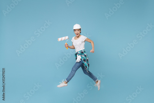 Side view funny young woman in protective helmet hardhat jump with paint roller isolated on blue background. Instruments accessories for renovation apartment room. Repair home concept. Looking camera.