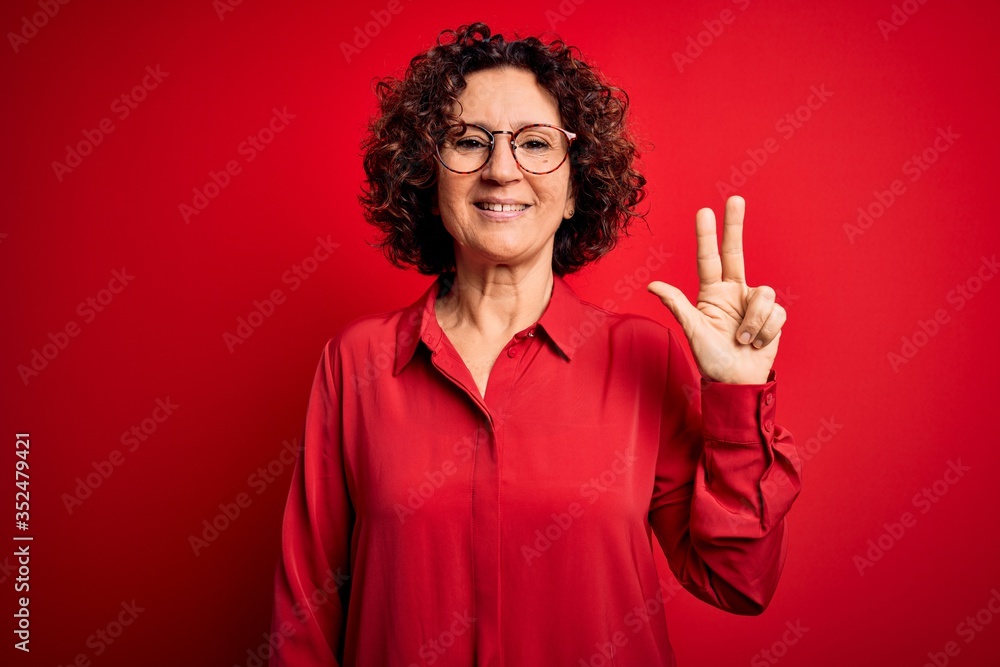 Middle age beautiful curly hair woman wearing casual shirt and glasses over red background showing and pointing up with fingers number three while smiling confident and happy. Middle age beautiful curly hair woman wearing casual shirt and glasses over red background showing and pointing up with fingers number three while smiling confident and happy.
