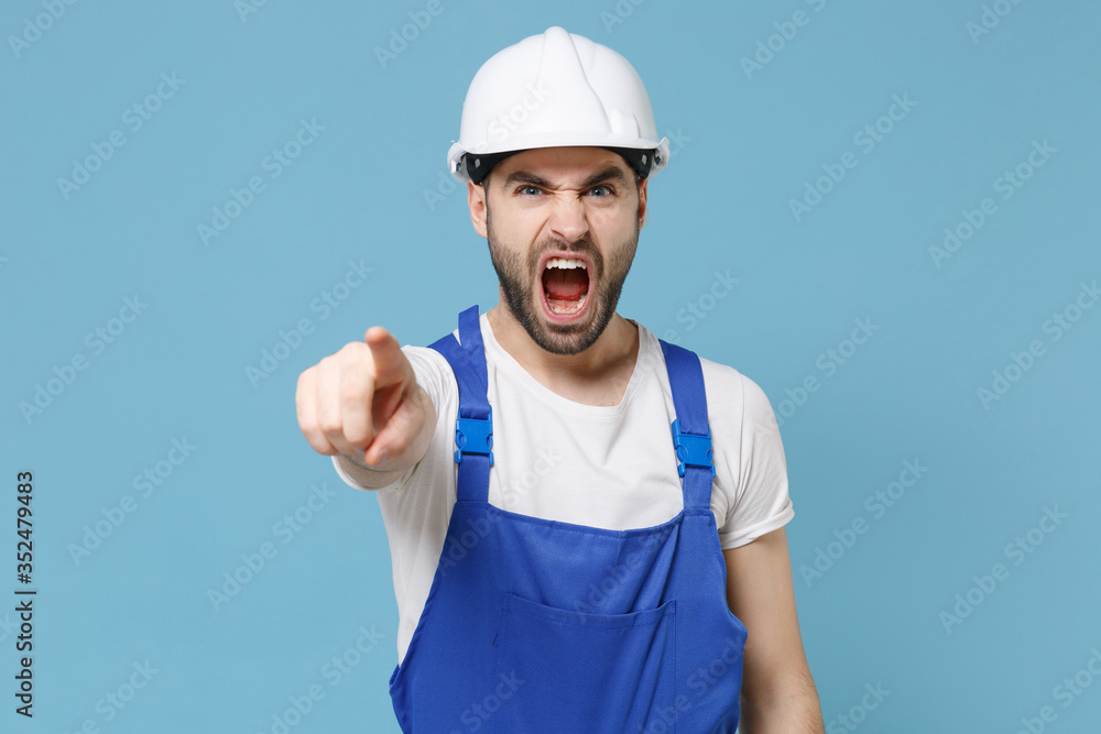 Angry man in coveralls protective helmet hardhat isolated on blue background. Instruments accessories for renovation apartment room. Repair home concept. Pointing finger on camera, screaming swearing. Angry man in coveralls protective helmet hardhat isolated on blue background. Instruments accessories for renovation apartment room. Repair home concept. Pointing finger on camera, screaming swearing.