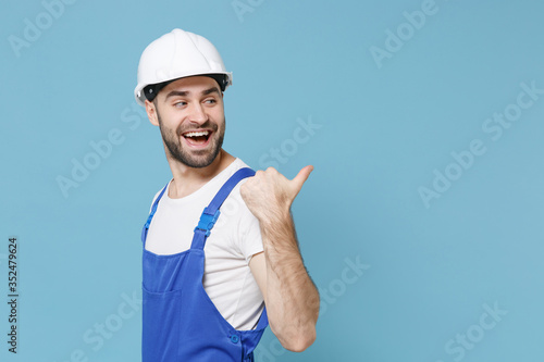 Side view of cheerful young man in coveralls protective helmet hardhat isolated on blue background. Instruments accessories for renovation apartment room. Repair home concept. Pointing thumb aside.