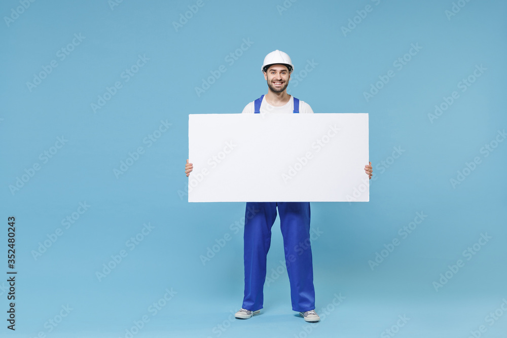 Smiling man in coveralls hardhat hold white empty blank billboard with place for text image isolated on pastel blue background. Instruments accessories renovation apartment room. Repair home concept. Smiling man in coveralls hardhat hold white empty blank billboard with place for text image isolated on pastel blue background. Instruments accessories renovation apartment room. Repair home concept.