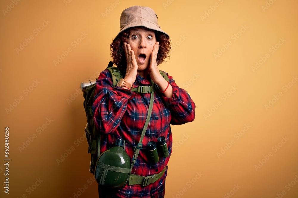 Middle age curly hair hiker woman hiking wearing backpack and water canteen using binoculars afraid and shocked, surprise and amazed expression with hands on face
