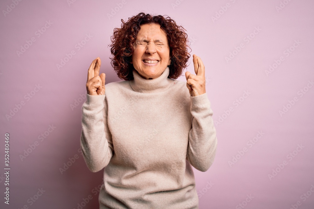 Middle age beautiful curly hair woman wearing casual turtleneck sweater over pink background gesturing finger crossed smiling with hope and eyes closed. Luck and superstitious concept. Middle age beautiful curly hair woman wearing casual turtleneck sweater over pink background gesturing finger crossed smiling with hope and eyes closed. Luck and superstitious concept.