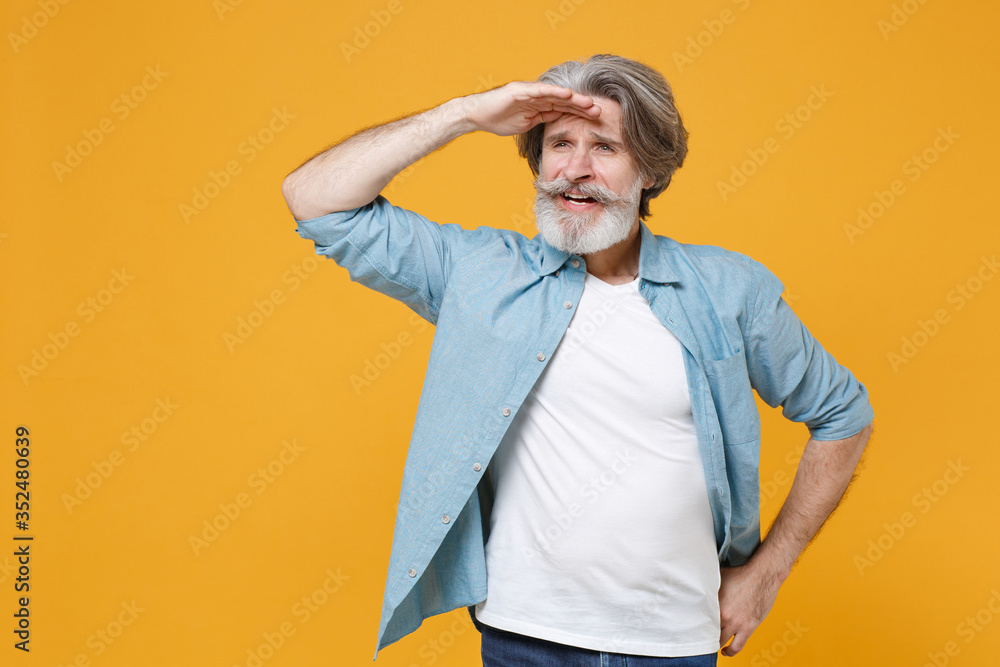 Elderly gray-haired mustache bearded man in casual blue shirt posing isolated on yellow background. People lifestyle concept. Mock up copy space. Holding hand at forehead looking far away distance. Elderly gray-haired mustache bearded man in casual blue shirt posing isolated on yellow background. People lifestyle concept. Mock up copy space. Holding hand at forehead looking far away distance.