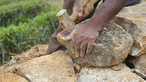 Male hands of indian stone worker cuts block of granite with a hammer for construction flowerbed. Adult unrecognizable stonemason hits a stone with sledgehammer in botanical garden. Slow motion