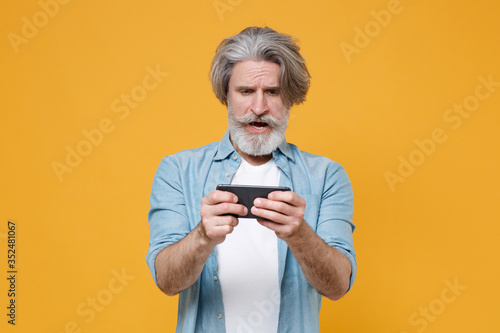 Perplexed elderly gray-haired mustache bearded man in casual blue shirt posing isolated on yellow background studio portrait. People lifestyle concept. Mock up copy space. Play game with mobile phone. © ViDi Studio