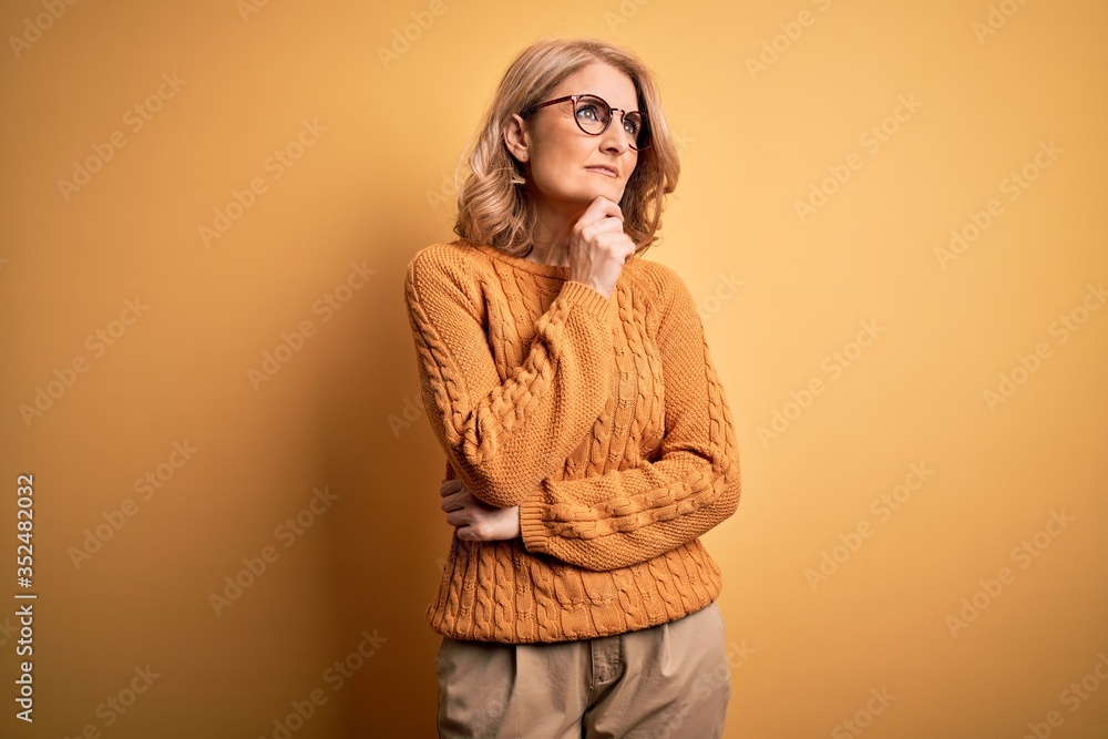 Middle age beautiful blonde woman wearing casual sweater and glasses over yellow background with hand on chin thinking about question, pensive expression. Smiling and thoughtful face. Doubt concept. Middle age beautiful blonde woman wearing casual sweater and glasses over yellow background with hand on chin thinking about question, pensive expression. Smiling and thoughtful face. Doubt concept.