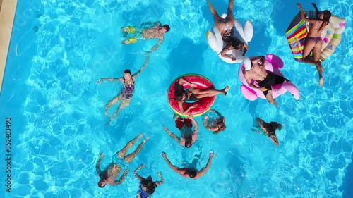 Aerial of friends having party in swimming pool with inflatable flamingo, swan, mattress. Happy young people relax at luxury resort on sunny day. View from above. Girls in bikini sunbathing in a sun.