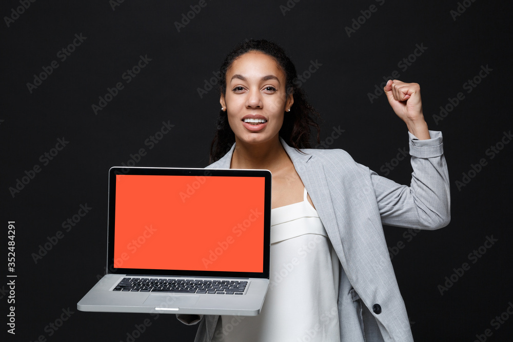 African american business woman in grey suit white shirt isolated on black background. Achievement career wealth business concept. Hold laptop pc computer with blank empty screen doing winner gesture. African american business woman in grey suit white shirt isolated on black background. Achievement career wealth business concept. Hold laptop pc computer with blank empty screen doing winner gesture.