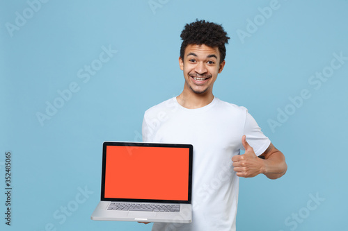 Smiling young african american guy in casual white t-shirt isolated on blue background. People lifestyle concept. Mock up copy space. Hold laptop pc computer with blank empty screen, showing thumb up.