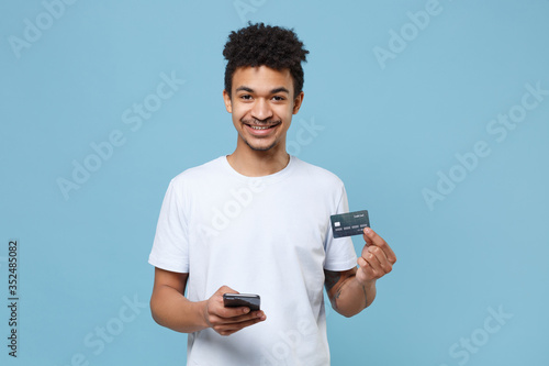 Smiling young african american guy in casual white t-shirt posing isolated on blue background studio portrait. People lifestyle concept. Mock up copy space. Using mobile phone hold credit bank card.