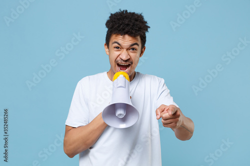 Nervous young african american guy in casual white t-shirt posing isolated on blue wall background. People lifestyle concept. Mock up copy space. Scream in megaphone, pointing index finger on camera.