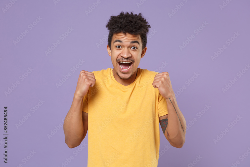 Excited young african american guy in casual yellow t-shirt posing isolated on pastel violet background studio portrait. People emotions lifestyle concept. Mock up copy space. Doing winner gesture. Excited young african american guy in casual yellow t-shirt posing isolated on pastel violet background studio portrait. People emotions lifestyle concept. Mock up copy space. Doing winner gesture.