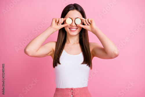 Photo of charming funny lady hold two macaroons cookies hiding eyes like specs playful mood wear white singlet dotted skirt isolated pastel pink color background © deagreez