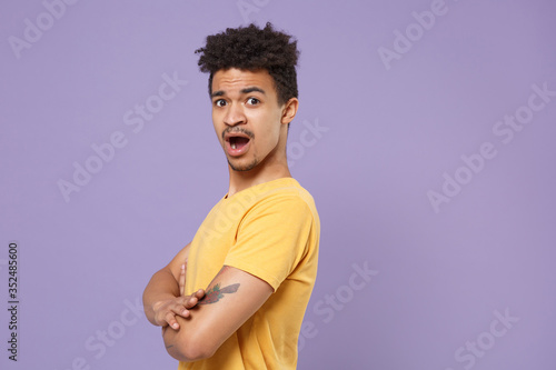 Side view of shocked young african american guy in casual yellow t-shirt posing isolated on pastel violet background. People lifestyle concept. Mock up copy space. Hold hands crossed, looking camera.