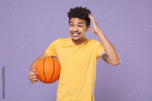 Excited young african american guy basketball player in yellow t-shirt isolated on violet wall background. People emotions, sport leisure lifestyle concept. Play basketball hold ball put hand on head.