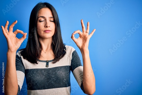 Young brunette woman with blue eyes wearing casual striped t-shirt standing over background relax and smiling with eyes closed doing meditation gesture with fingers. Yoga concept.