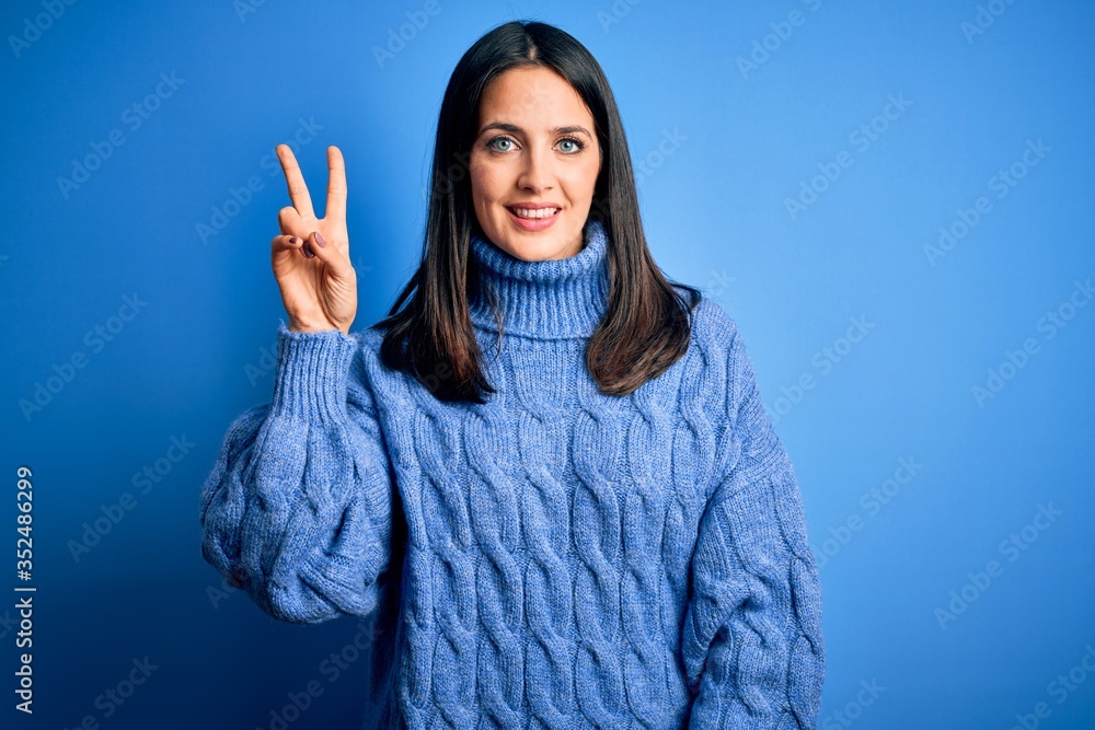 Young brunette woman with blue eyes wearing casual turtleneck sweater smiling looking to the camera showing fingers doing victory sign. Number two. Young brunette woman with blue eyes wearing casual turtleneck sweater smiling looking to the camera showing fingers doing victory sign. Number two.