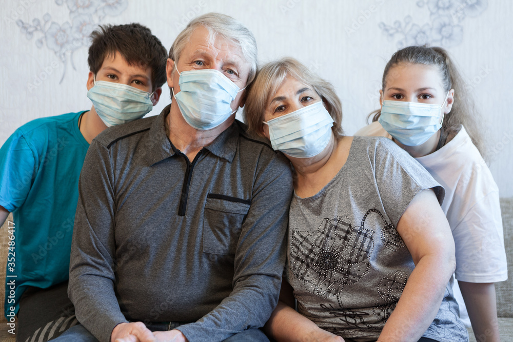 Four people portrait, grandparents with their teen age grandchildren wearing facial masks, sitting on sofa in a domestic room