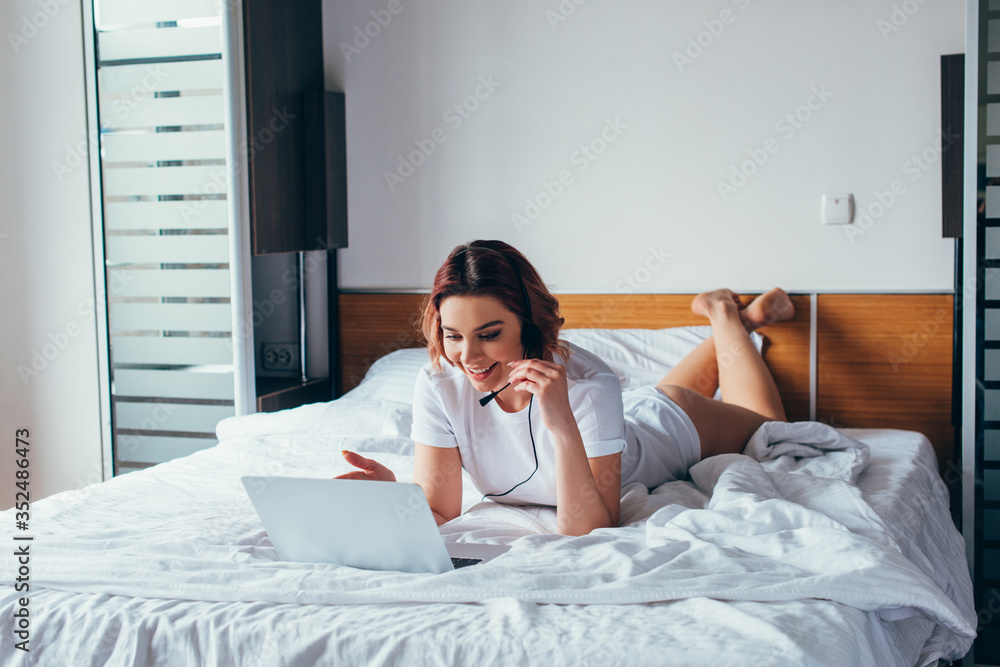 smiling girl making video call with headset and laptop in bed during self isolation smiling girl making video call with headset and laptop in bed during self isolation