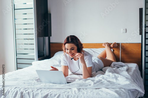 smiling girl making video call with headset and laptop in bed during self isolation