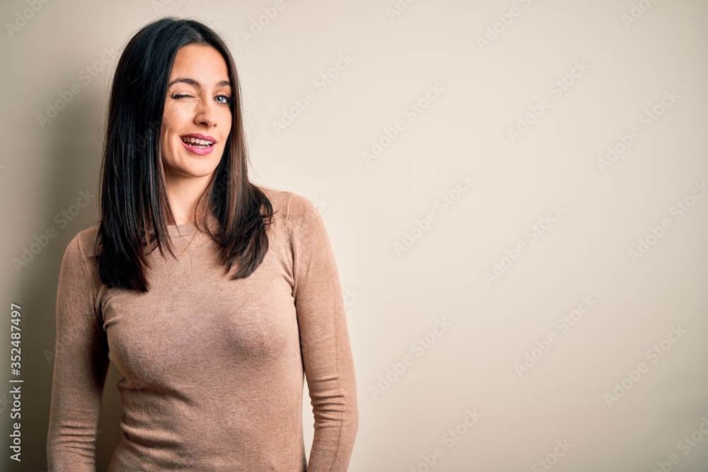 Obraz premium Young brunette woman with blue eyes wearing casual sweater over isolated white background winking looking at the camera with sexy expression, cheerful and happy face.