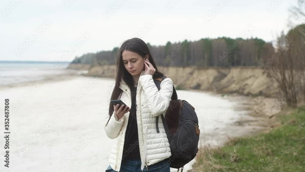 young girl is traveling in nature with Hiking backpack on back. girl is standing on edge of cliff enjoying relaxation and the landscape turns on her smartphone and puts on headphones. young girl is traveling in nature with Hiking backpack on back. girl is standing on edge of cliff enjoying relaxation and the landscape turns on her smartphone and puts on headphones.