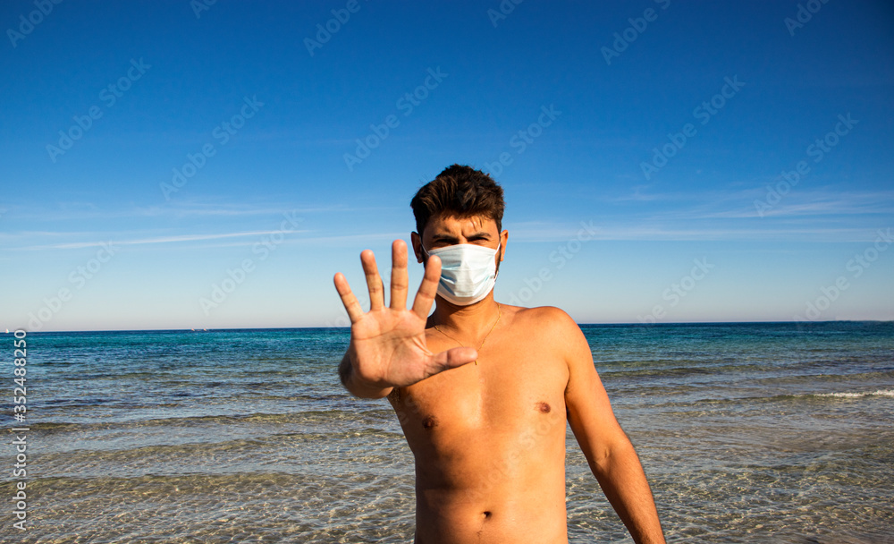 Masked man on the beach makes stopping hand gesture. Coronavirus concept.
