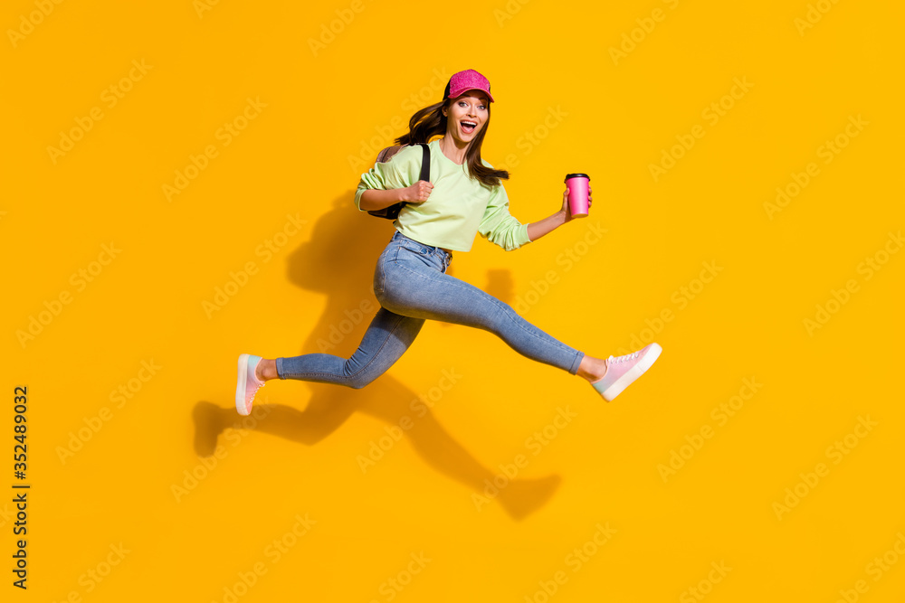 Full body profile photo of energetic lady student hold backpack takeout coffee jump high rushing lessons wear green cropped sweatshirt jeans shoes cap isolated vivid yellow color background