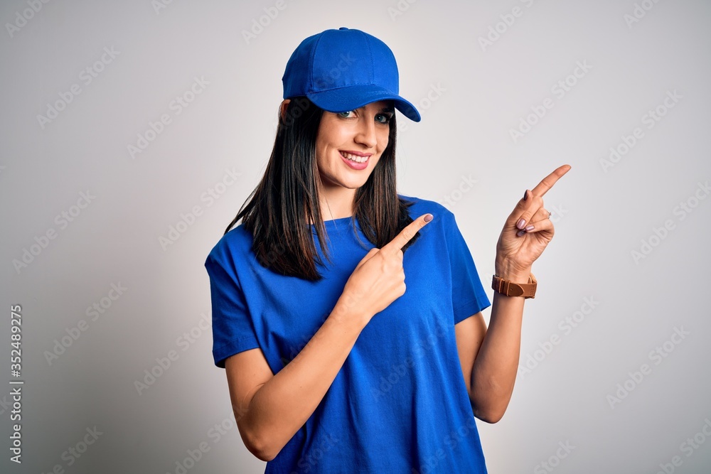 Young delivery woman with blue eyes wearing cap standing over blue background smiling and looking at the camera pointing with two hands and fingers to the side.