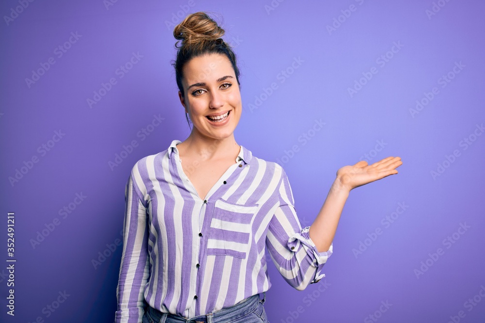 Young beautiful blonde woman wearing casual striped shirt standing over purple background smiling cheerful presenting and pointing with palm of hand looking at the camera. Young beautiful blonde woman wearing casual striped shirt standing over purple background smiling cheerful presenting and pointing with palm of hand looking at the camera.