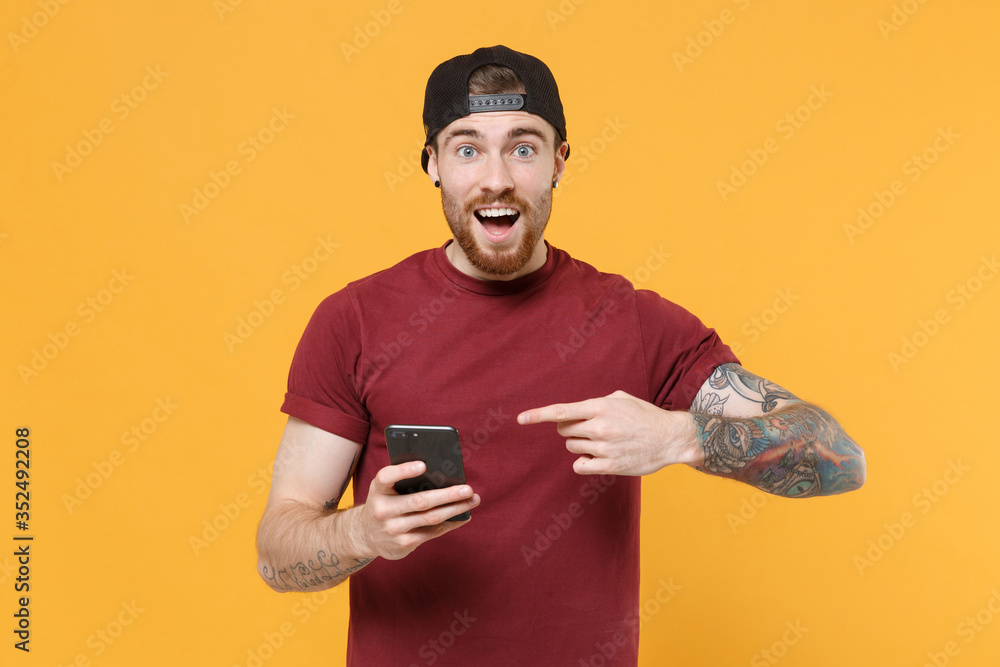Amazed young bearded tattooed man guy in casual t-shirt black cap posing isolated on yellow wall background studio. People lifestyle concept. Mock up copy space. Pointing index finger on mobile phone. Amazed young bearded tattooed man guy in casual t-shirt black cap posing isolated on yellow wall background studio. People lifestyle concept. Mock up copy space. Pointing index finger on mobile phone.